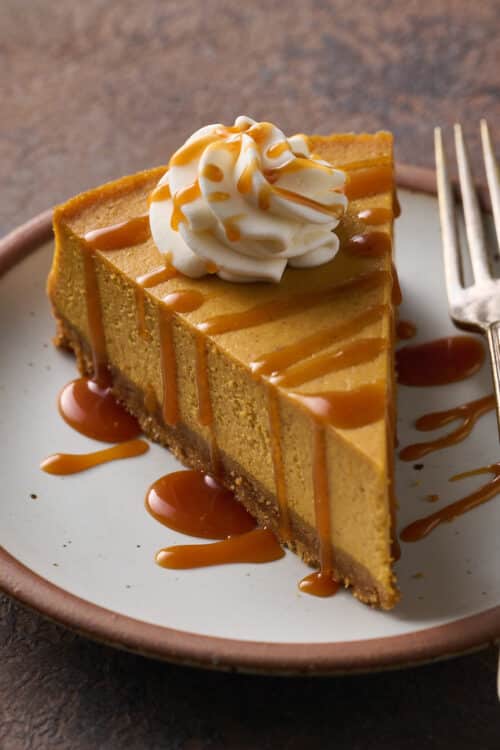 A slice of pumpkin cheesecake on a plate topped with whipped cream and salted caramel sauce.